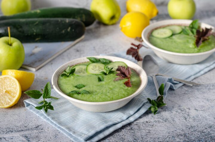 Grüne Gurken-Gazpacho im Teller. Auf einem Tisch dekoriert mit Gurken, Äpfeln und Zitronen