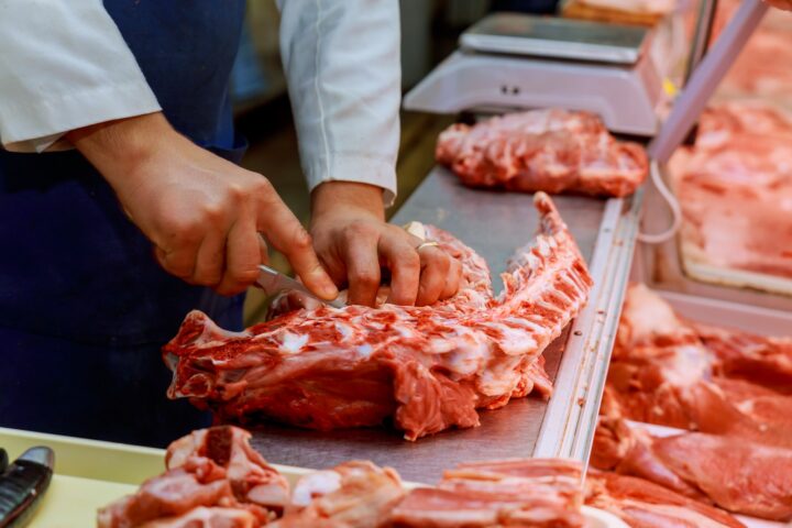 Hands of a butcher cutting slices of raw meat with knife at counter in shop