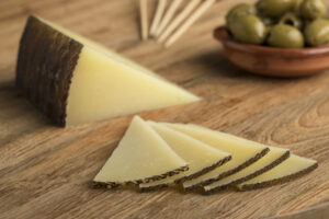 Scheiben von Manchego Käse auf einem Holzbrett mit grünen Oliven im Hintergrund